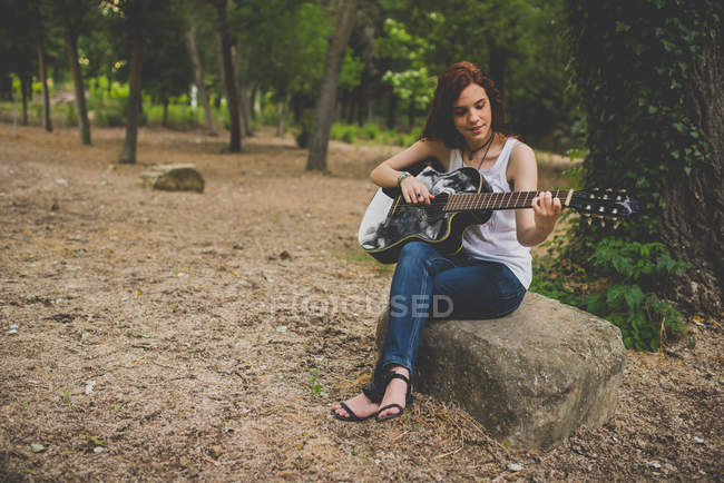 Retrato de menina sardenta sorridente sentado em pedra na floresta e tocando guitarra — Fotografia de Stock
