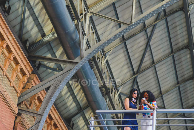 Низький кут зору двох жінок, що спираються на перила балкона і використовують мобільний телефон під залізним дахом . — стокове фото