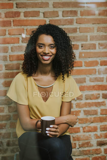 Mujer morena sonriente con taza mirando a la cámara en el fondo de la pared de ladrillo. Tiro vertical en interiores - foto de stock