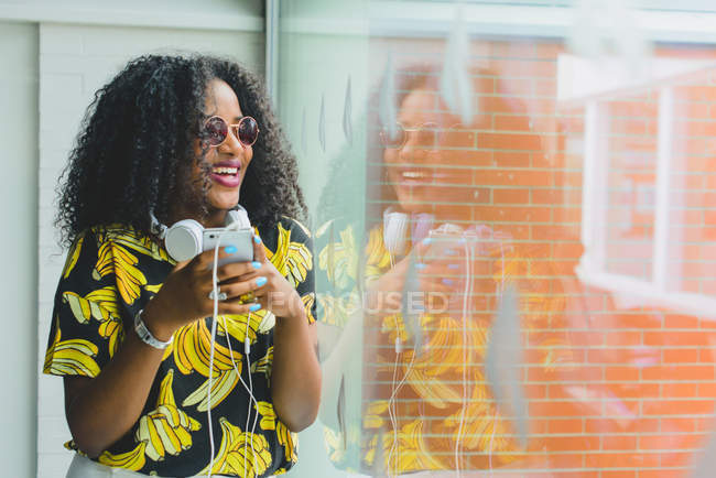 Portrait de fille souriante dans des lunettes de soleil en utilisant un smartphone et en regardant la fenêtre — Photo de stock