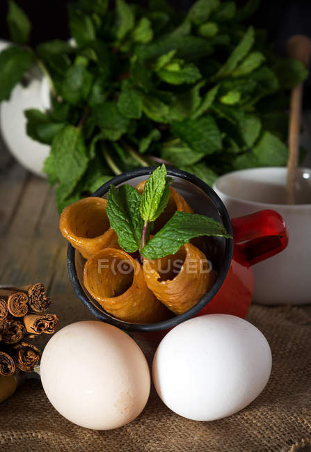Close up view of mug with sweet dough tubes and mint leaves lying on sacking with eggs — Stock Photo