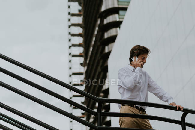 Vista de bajo ángulo del hombre de negocios en camisa blanca de pie en el pasillo de las escaleras y hablando en el teléfono inteligente sobre edificios de negocios fachada de vidrio - foto de stock