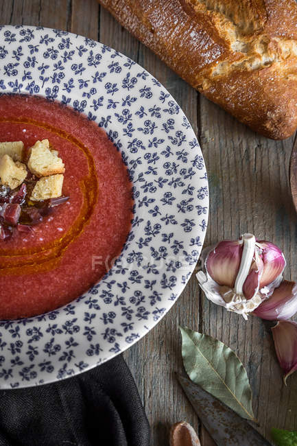 Sopa de tomate em prato estampado em mesa de madeira com pão e ingredientes — Fotografia de Stock