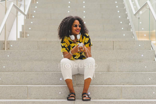 Porträt einer fröhlichen Frau mit Kopfhörern um den Hals, die ihr Smartphone benutzt und wegschaut, während sie auf der Treppe sitzt — Stockfoto