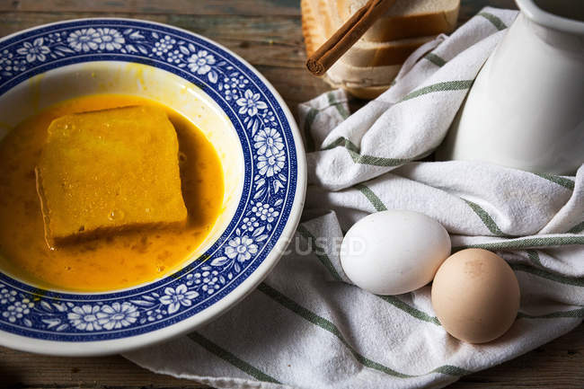 Высокий угол обзора декоративной тарелки с ломтиком хлеба, лежащим в разбитом яйце на столе с полотенцем и яйцами — стоковое фото
