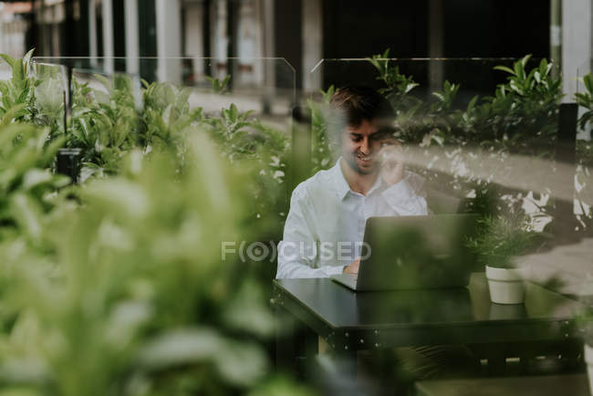 Portrait of smiling businessman sitting at cafe terrace table with laptop and talking on smartphone — Stock Photo