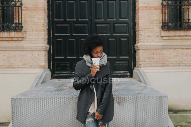 Портрет девушки в пальто позирует с чашкой кофе над фасадом здания — стоковое фото