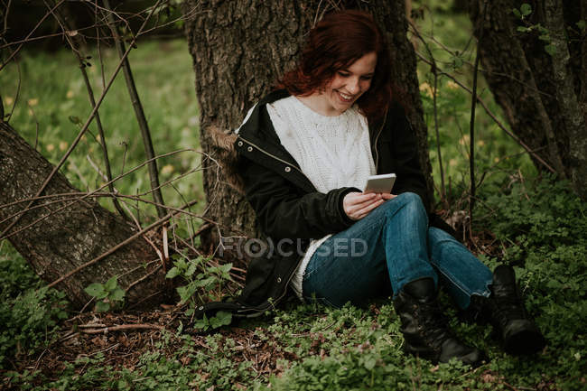 Улыбающаяся девочка сидит у дерева и просматривает смартфон — стоковое фото
