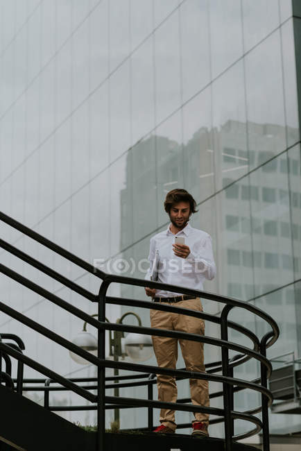 Portrait of smiling businessman in white shirt standing on stairs passage and using smartphone  over business building glass facade on background — Stock Photo