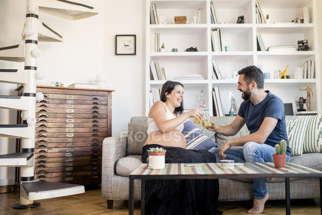 Father and pregnant mother cheering, sitting on sofa and drinking some juice. — Stock Photo