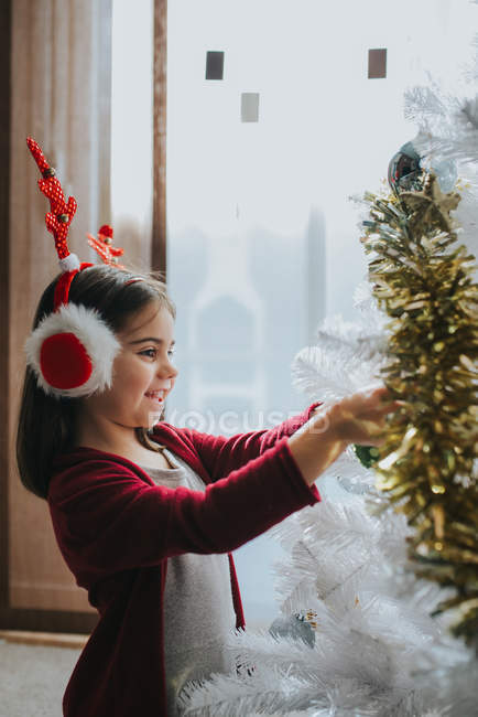 Side view of smiling girl wearing fur earmuffs with antlers placing baubles on decorative Christmas tree — Stock Photo