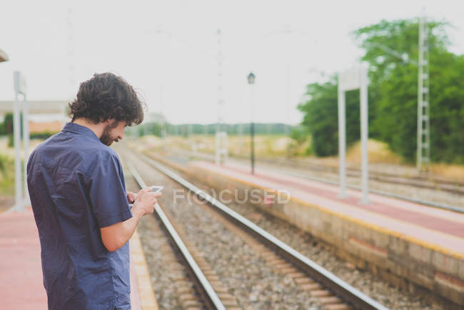 Вид сзади на человека с помощью смартфона на железнодорожной платформе в сельской местности — стоковое фото