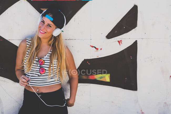 Retrato de una atractiva joven rubia escuchando música en auriculares contra la pared de graffiti. - foto de stock