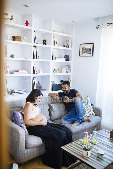 Jovem pai e mãe grávida esperando para o parto, sentado no sofá, segurando câmera fotográfica. — Fotografia de Stock