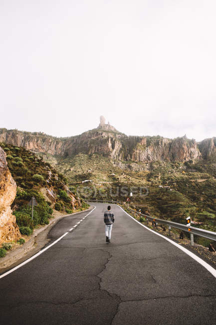 Вид сзади на человека, идущего по асфальтированной дороге по горной местности — стоковое фото