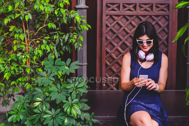 Retrato de menina com fones de ouvido em torno do pescoço sentado perto de plantas envasadas e usando smartphone — Fotografia de Stock