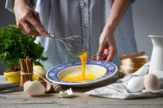 Sección media de hembra batiendo huevos con batidor en plato a la mesa con ingredientes - foto de stock