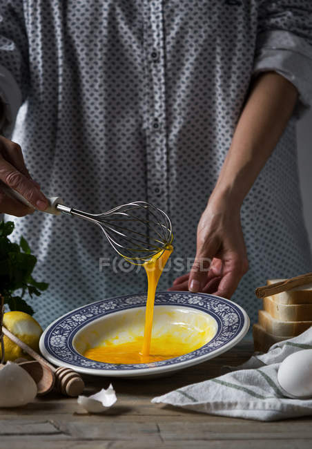 Sección media de hembra batiendo huevos con batidor en plato a la mesa con ingredientes y utensilios de cocina - foto de stock