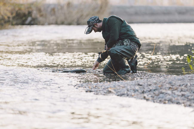 Vista lateral del hombre arrodillado por el río y la pesca con caña - foto de stock