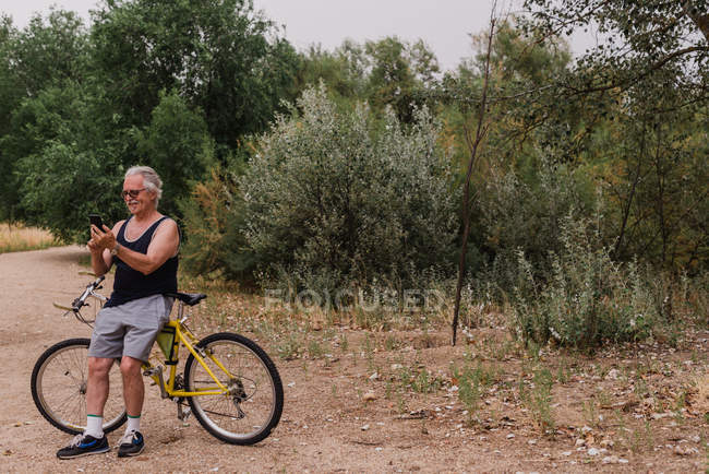 Портрет старшого чоловіка, що спирається на велосипед і перегляд смартфона в парку — стокове фото