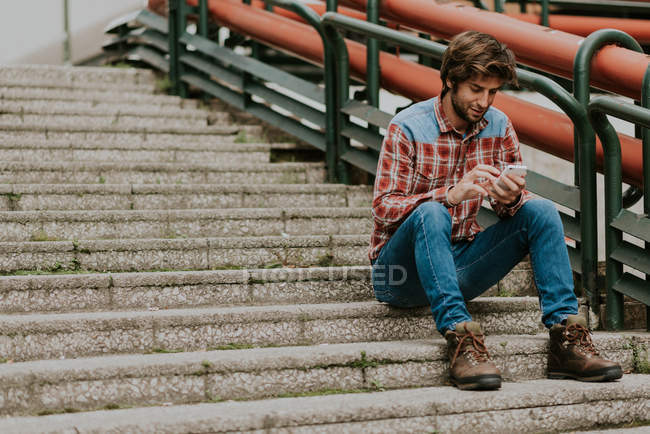 Ritratto di uomo barbuto sorridente in abiti casual seduto sui gradini della strada e utilizzando il telefono — Foto stock