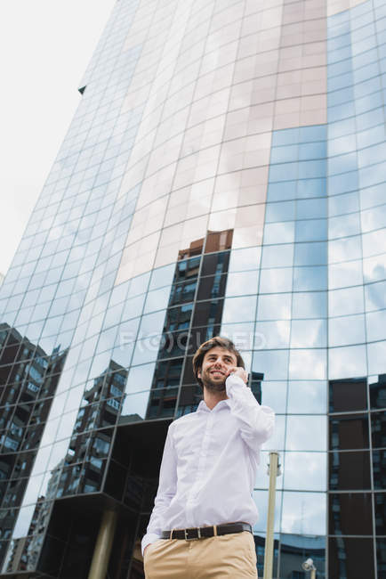 Retrato de baixo ângulo de homem de negócios sorridente em camisa branca falando em smartphone sobre fachada de prédio de negócios no centro da cidade cena urbana — Fotografia de Stock
