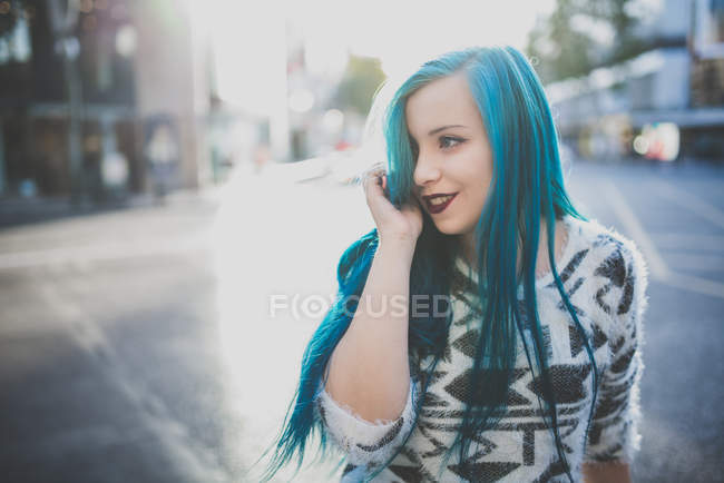 Retrato de menina vestindo suéter macio alisando seu cabelo liso azul e olhando para o lado na cena da rua — Fotografia de Stock