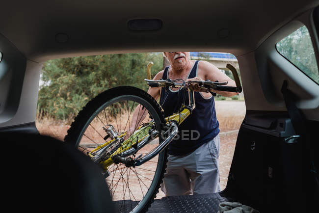Старший чоловік кладе велосипед у багажник автомобіля на сільській місцевості — стокове фото