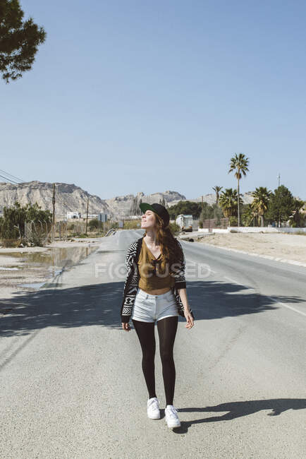 Full length shot of young stylish woman walking down street in sunlight on background of rocky hills. — Stock Photo