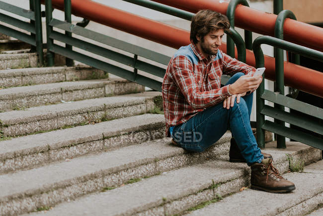 Vista lateral del hombre barbudo con ropa casual sentado en las escaleras de la calle y utilizando el teléfono inteligente - foto de stock
