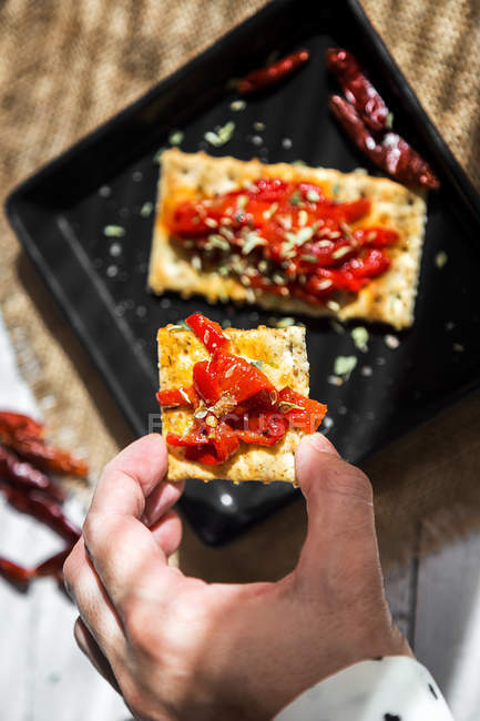 Above view of hand holding cracker with dried tomatoes ove baking pan on table — Stock Photo