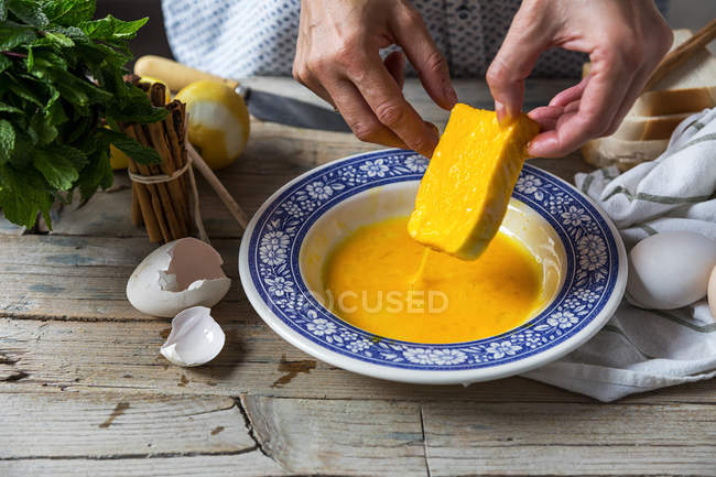 Крупним планом вид жіночих рук, що стукають хліб скибочками в тарілці з розбитими яйцями на сільському дерев'яному столі — стокове фото