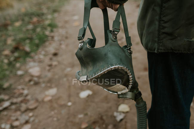 Cropped image of gas mask hanging in male hand over rural road — Stock Photo