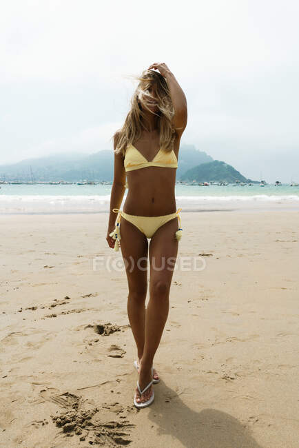 Young beautiful woman with slim body posing in yellow swimsuit covering face with hair on beach. — Stock Photo