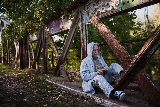 Old man in hoodie sitting on metal bridge construction and looking at something in his hands. Copyspace — Stock Photo