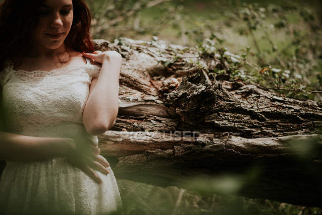 Обрізати дівчину в білій сукні, спираючись на дерево в лісі — стокове фото