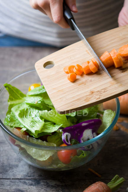 Руки на полях добавляют морковные ломтики из доски в миску с салатом — стоковое фото
