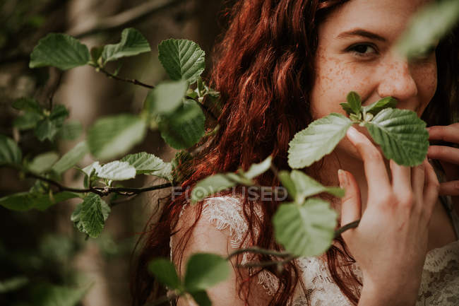 Retrato de chica pelirroja mirando a la cámara a través de hojas verdes - foto de stock