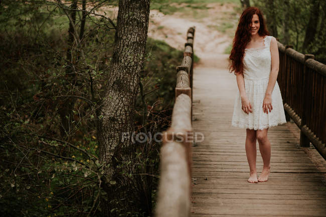 Веселая рыжая девушка позирует в белом платье на деревянном мосту — стоковое фото