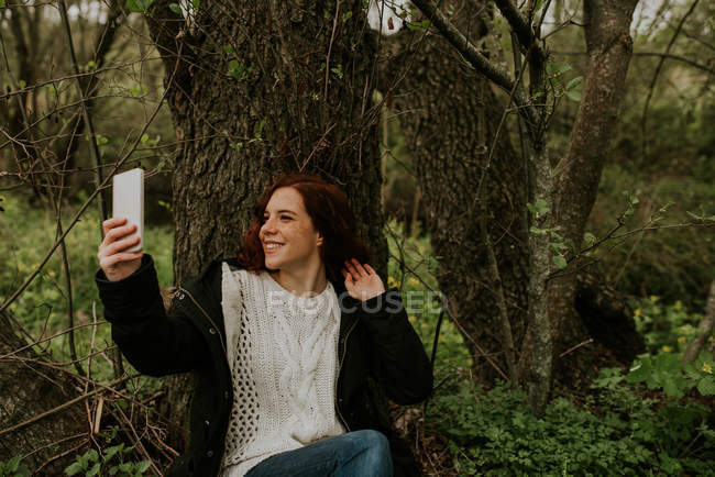 Усміхнена дівчина сидить на дереві і приймає селфі зі смартфоном — стокове фото