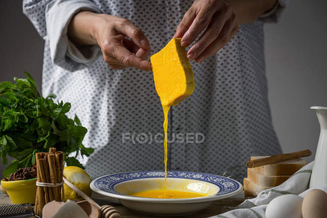 Середина жінки тримає скибочку хліба, поливаючи розбиті яйця в тарілці на кухонному столі — стокове фото