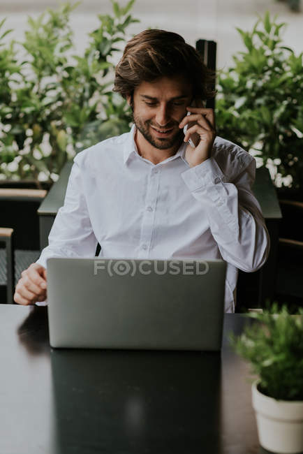 Портрет усміхненого бізнесмена, який сидить за столом і розмовляє через смартфон, використовуючи ноутбук на терасі кафе — стокове фото