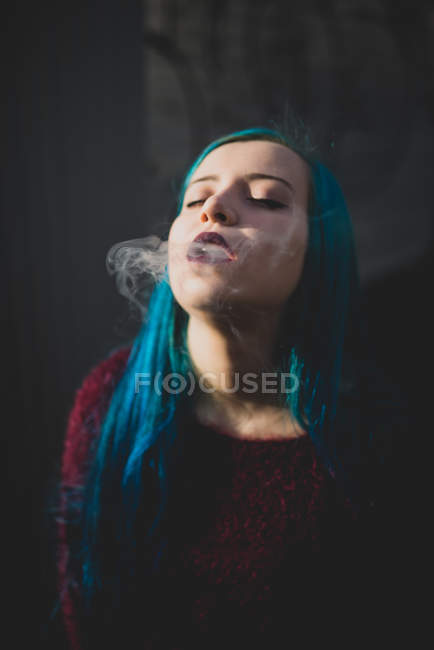 Portrait of blue haired girl posing at camera and smoking out cigarette smoke — Stock Photo