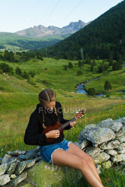 Mulher sentada na cerca de pedra rural na natureza e jogando ukulele — Fotografia de Stock