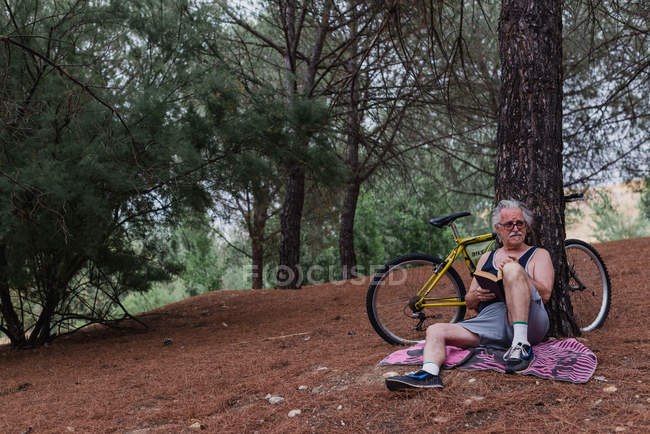 Senior sentado en el suelo con libro en las manos y apoyado en el árbol con bicicleta estacionada en el bosque - foto de stock