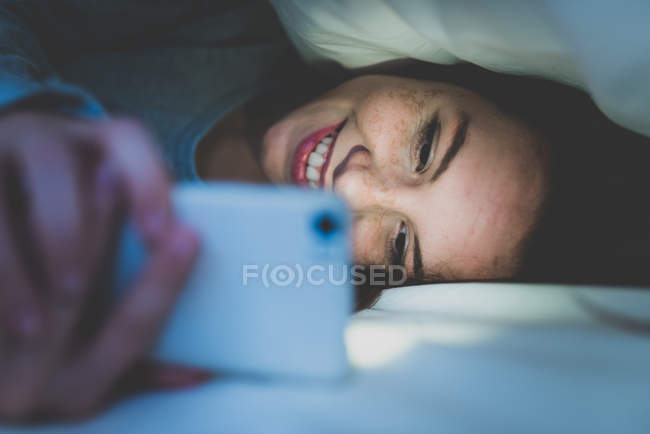 Menina deitada na cama sob travesseiro e usando smartphone — Fotografia de Stock