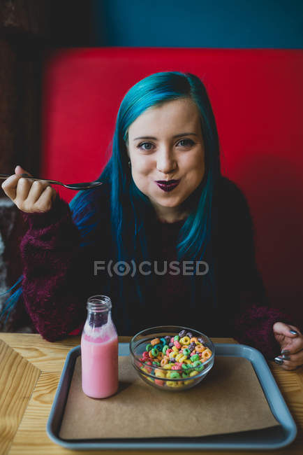 Retrato de menina de cabelos azuis sentada à mesa do café, comendo cereais com colher e olhando para a câmera — Fotografia de Stock
