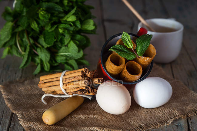 Still life of fried honey dough tubes with mint in mug lying on sacking with eggs over rural wooden table — Stock Photo