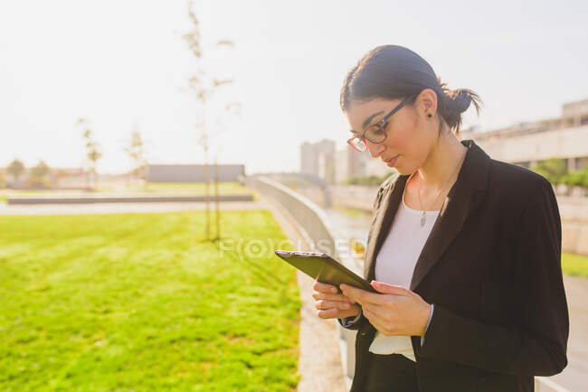 Vista lateral de mujer joven en gafas mirando su tableta mientras está de pie al aire libre. Espacio de copia - foto de stock