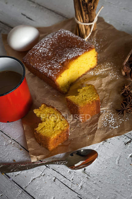Vista de ángulo alto de rodajas caseras de pastel de limón en papel de panadería con ingredientes y taza roja en mesa de madera rural - foto de stock